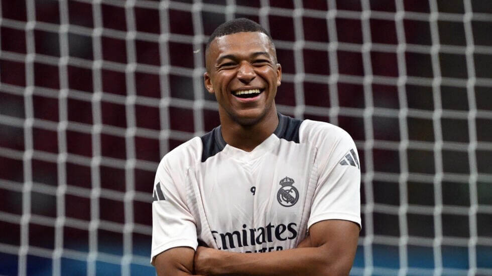 Kylian Mbappe, in white Emirates uniform, smiles at the crowd just in front of the net during a match. Mbappe demands unpaid wages to be paid to him by PSG.