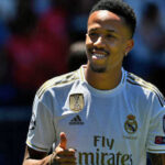 Eder Militao, in white uniform, flashes a sign of approval during a match.