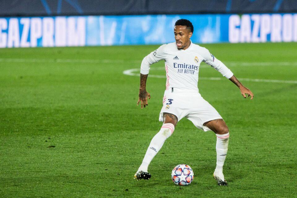 Eder Militao, in white uniform, protects the ball as he ponder his next move in a match