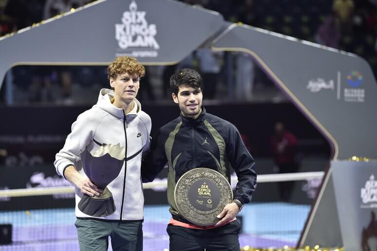 Jannik Sinner, in white jacket, and Carlos Alcaraz, in black, share the stage while holding their trophies. Sinner tops Alcaraz for Six Kings Slam title.