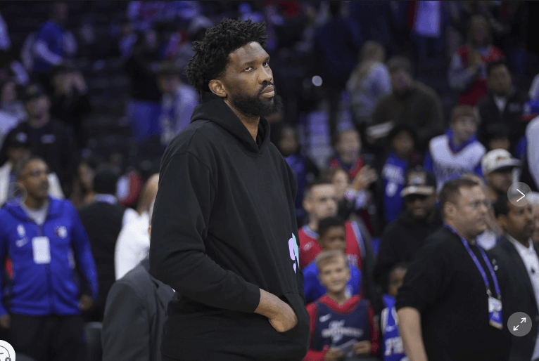 Sixers center Joel Embiid, in black sweat shirt and pants, looks on from the sidelines. Joel Embiid gets a three-game suspension from the NBA for shoving a columnist after the Philadelphia 76ers match over the weekend.